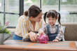 © thepoo - Asian mother and little daughter putting coins into piggy bank.