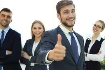 Wall Mural - Feeling confident in his team. Happy businessman showing his thumb up and smiling while his colleagues standing in the background.