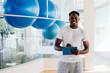 © twinsterphoto - Handsome African American personal trainer standing with clipboard by fitness balls, smiling and looking at camera