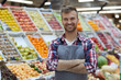 Waist up portrait of handsome young man working in supermarket and smiling at camera while posing by fruit stand, copy space