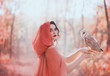 mystical pagan woman with covered head in peach scarf in forest, holds cute little barn owl. lady with dark curled hair, bright make-up and fair pale skin, daughter of autumn forest caring for bird