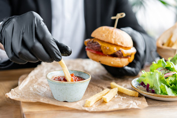 Young man in business suit and black gloves eating delicious juicy cheeseburger with beef chop. Burgers in the cafe with salad and fries, fast food.