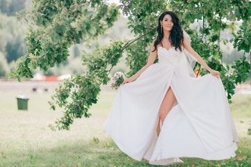 Happy tanned bride in beautiful white dress posing with boquet of flowers in front of tree in summer day.