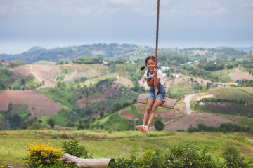 Wall Mural - Happy asian child girl having fun to play on wooden swings in playground with beautiful nature