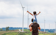 Father And Daughter Having Fun To Play Together. Asian Child Girl Riding On Father's Shoulders In The Wind Turbine Field