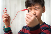 Closeup Of Smart Preteen / Teenage Asian Boy's Face And Hand  Holding Black And Red Probe Of Multimeter Connects With Red Wire, Checking Electric Current. STEM Education And Learning By Doing Concept.