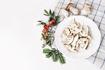 Wall Mural - Autumn food arrangement. Composition of whole and sliced porcino mushrooms, ceps on plate. Rowan berries, leaves, fir branches and checkered tea towel on white table background. Fall design, flat lay