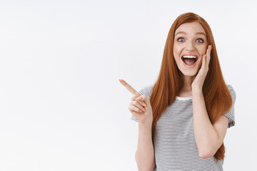 Wall Mural - Cheerful charismatic impressed redhead young girl open mouth smiling astonished pointing upper left corner reacting awesome cool news, standing white background thrilled, fan finally come concert