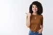 © Cookie Studio - Have you met my friend. Glad good-looking confident stylish female curly dark hair pointing thumb left introduce product smiling happily recommend good promo hold hand jeans pockets assured