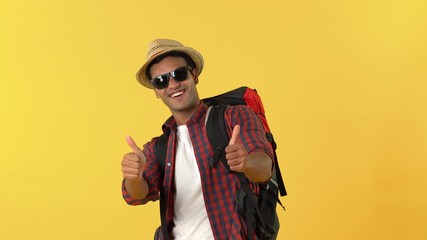 Wall Mural - Portrait of handsome Indian tourist man wearing sunglasses saluting and showing thumbs up sign in front of camera against yellow background