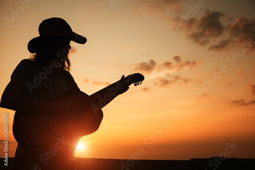 Plakaty Country & Western  sylwetka-mlodej-kobiety-grajacej-na-gitarze-o-zachodzie-slonca