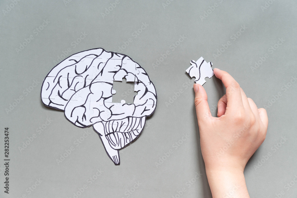 Female hand trying to connect a missing jigsaw puzzle of human brain on gray background. Creative idea for solving problem, memory loss, dementia or Alzheimer's disease concept. Mental health care. - obrazy, fototapety, plakaty 