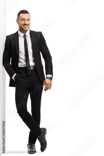 Cuadros en Lienzo Young cheerful man in a black suit leaning on a wall
