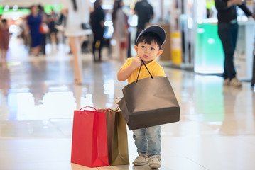 Kid shopping. Asian little boy in yellow shirt and jeans enjoyment searching something toys in his hand shopping bags while standing in the shopping mall.