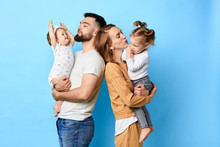 Two Caring Parents Standing Back To Back And Holding Their Children In Arms. Close Up Photo. Isolated Blue Background. Studio Shot.