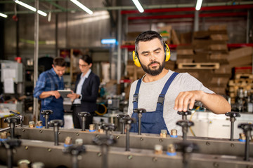 Young bearded mechanic in protective headphones repairing industrial machine