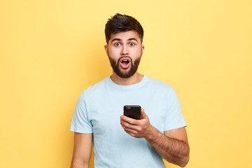 handsome emotional man with shocked expression holding smart phone looking at the camera. People, emotions and technology concept.close up portrait, isolated yellow background, studio shot