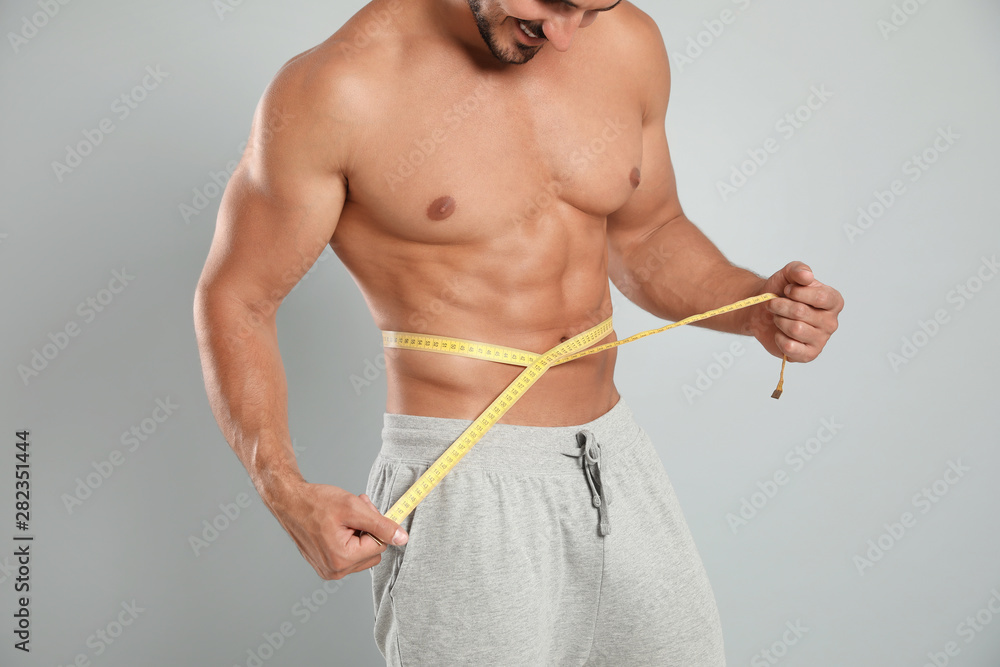 Young man with slim body using measuring tape on grey background, closeup view - obrazy, fototapety, plakaty 