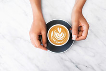 Woman Hands Holding Gray Cup Of Fresh Cappuccino With Classic Latte Art On White Marble Table Trendy Background. Empty Place For Text, Copy Space. Coffee Addiction Concept. Top View.