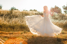 Bride On A Summer Field In White Wedding Dress Rolling And Dancing In Sunset Light. Sun Beams Seen Through Transparent Dress Skirt Fabric. Rustic Or Boho Outdoor Wedding Concept. Selective Soft Focus.