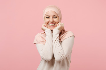 Poster - Joyful young arabian muslim woman in hijab light clothes posing isolated on pink background studio portrait. People religious Islam lifestyle concept. Mock up copy space. Keeping hands near cheeks.