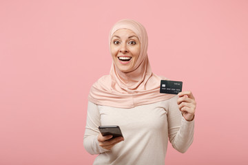 Excited young arabian muslim woman in hijab light clothes posing isolated on pink background. People religious Islam lifestyle concept. Mock up copy space. Using mobile phone, hold credit bank card.