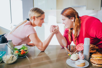 Dilemma. To lose weight or not. Fitness vs body lotion. Athletic slender young woman contends in armwresling with fat body positive plus size model. Serious fight between two women.