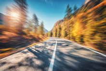Mountain Road In Autumn Forest At Sunset With Motion Blur Effect.  Asphalt Road And Blurred Background With Orange Trees, Blue Sky With Sun In Fall. Fast Driving. Beautiful Highway. Transportation