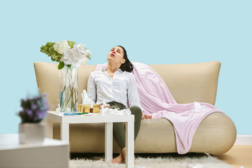 Wall Mural - Young woman suffering from hausehold dust or seasonal allergy. Sneezing in the napkin and sitting surrounded by used napkins on the floor and sofa. Taking medicines with no result. Healthcare concept.