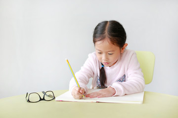 Portrait of smiling little Asian girl writes in a book or notebook with pencil on table in classroom against white background.