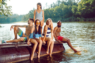 Wall Mural - Group of happy friends having fun while splashing water and laughting on the pier on river. Joyful women and men in swimsuit at riverside in sunny day. Summertime, friendship, resort, relations