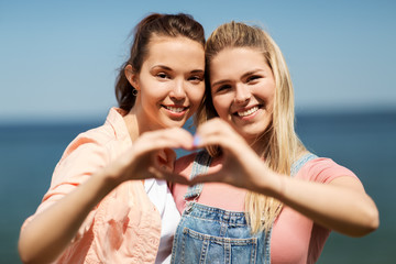 Poster - leisure and friendship concept - happy smiling teenage girls or best friends at seaside in summer making hand heart gesture