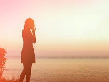 Silhouette Of A Young Woman Who Stands On A Rock In The Sea And Looks At The Sunset. Focus On The Girl