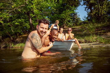 Wall Mural - Happy group of friends having fun while laughting and swimming in river. Joyful men and women in swimsuit in a boat at riverside in sunny day. Summertime, friendship, resort, weekend concept.