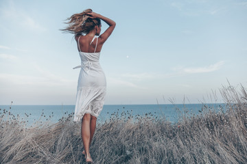 Young woman in white dress back view of staying on the cliff looking forward at amazing ocean view. Free space for text. Travel and enjoying life concept