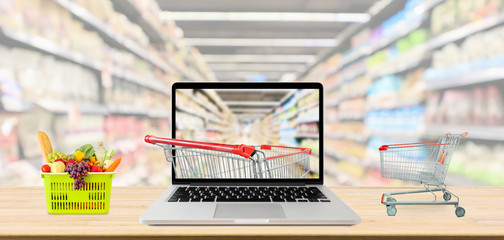 Wall Mural - supermarket aisle blurred background with laptop computer and shopping cart on wood table grocery online concept