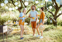 Young Friends Having Fun, Standing Together With Drinks In The Beautifully Decorated Backyard Or Garden During A Festive Lunch Or Party