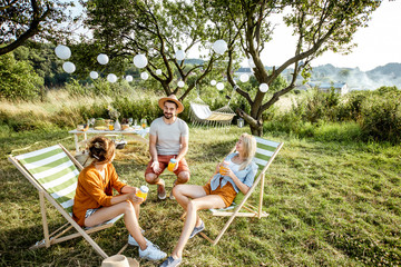 Young and cheerful friends relaxing with drinks, sitting on the sunbeds in the beautifully decorated backyard or garden during a festive meeting