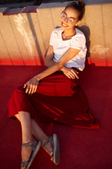 Wall Mural - Bright summer lifestyle portrait of young pretty woman in eyewear, red skirt and white T-shirt, lying on a bright red floor outdoors.