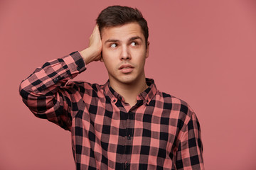 Wall Mural - Portrait of young pencive man in checkered shirt, looks away, stands over pink background and touches head, looks tired and sad.