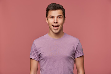 Wall Mural - Portrait of handsome cheerful guy wears in blank t-shirt, looks at the camera with happy amazed expression, stands over pink background.