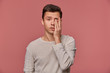 © timtimphoto - Portrait of young attractive man in white long sleeve, looks at the camera and touches face, stands over pink background, looks tired and sad.