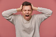 © timtimphoto - Close up of young attractive man in blank longs leeve, stands over pink background with closed eyes and screaming, looks angry and unhappy, touches head.