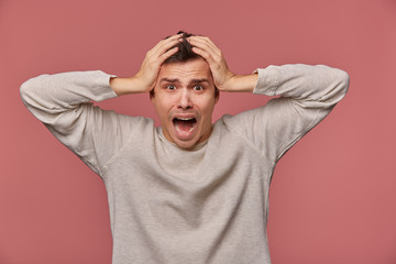 Wall Mural - Young scared man in blank long sleeve, hears very bad news, holds head and looks at the camera with shocked expression, stands over pink background and screaming, looks afraid.