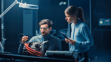 Male And Female Automotive Engineers With A Tablet Computer And Inspection Tools Are Having A Conversation While Testing An Electric Engine In A High Tech Laboratory With A Concept Car Chassis.