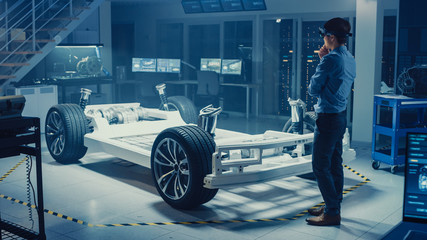 Automotive Engineer Working on Electric Car Chassis Platform, Using Augmented Reality Headset. In Innovation Laboratory Facility Concept Vehicle Frame Includes Wheels, Suspension, Engine and Battery.