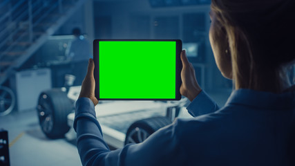 Female Engineer Holds a Tablet Computer with Green Screen Mock Up and Looks at Electric Car Chassis Prototype with Wheels, Batteries and Engine Standing in a High Tech Development Laboratory.