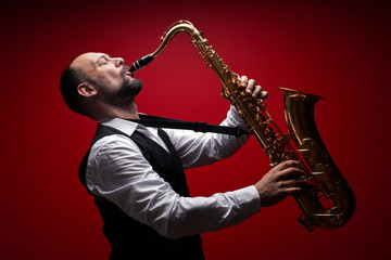 Portrait of professional musician saxophonist man in  suit plays jazz music on saxophone, red background in a photo studio