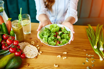 Wall Mural - Young happy woman eating salad in the beautiful interior with green flowers on the background and fresh ingredients on the table. Healthy food concept