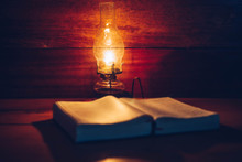 Close Up Of Oil Lamp With Blurred Holy Bible On Wood Table In Dark Room, Bible Study Or Vintage Book Concept